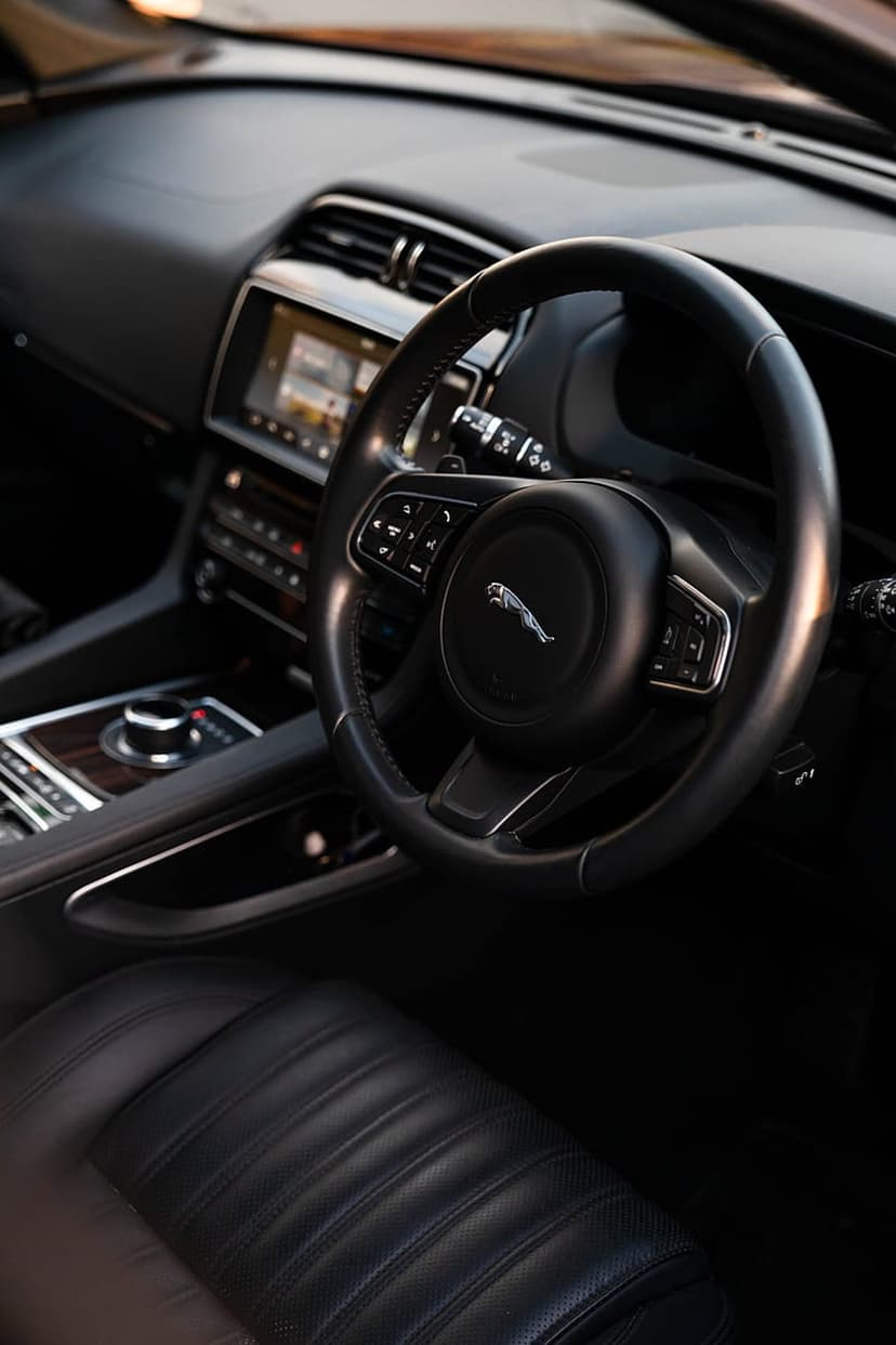 Jaguar steering wheel with chrome emblem sits before a glowing dashboard and quilted leather seat in a car cabin.