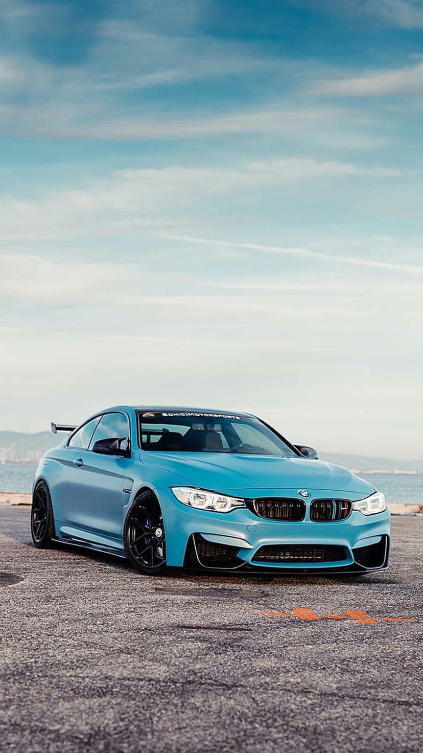 Blue BMW M4 coupe with a rear spoiler parked on asphalt facing a calm ocean under a hazy, clouded sky.