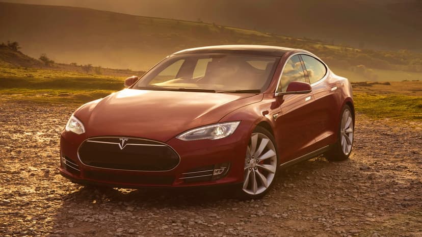 Red Tesla Model S on a gravel path during golden hour with rolling misty hills in the scenic background.