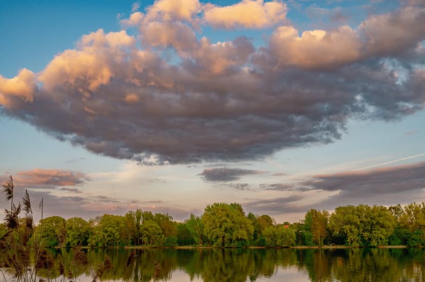Sunset clouds in shades of orange and pink reflect on a mirror-like lake bordered by a dark silhouette of trees.