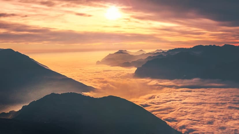 Snow-dusted mountain peaks glow orange as they emerge from a thick white sea of clouds during an alpine sunrise.