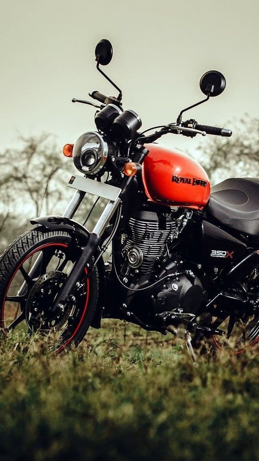 Royal Enfield Meteor 350X orange motorcycle with chrome details parked in a lush green field under a soft sky.