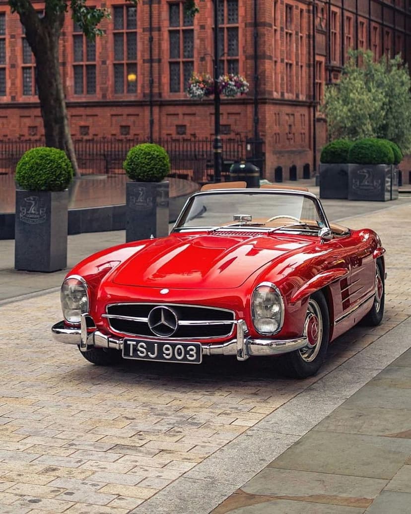 Red Mercedes 300 SL Roadster with tan interior parked on a cobblestone street near brick buildings and greenery.