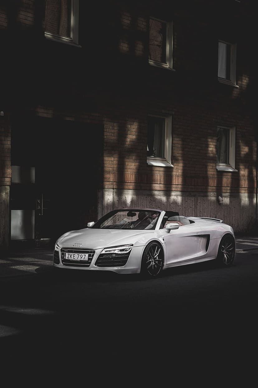White Audi R8 convertible parked on a street with harsh shadows against a red brick building exterior.