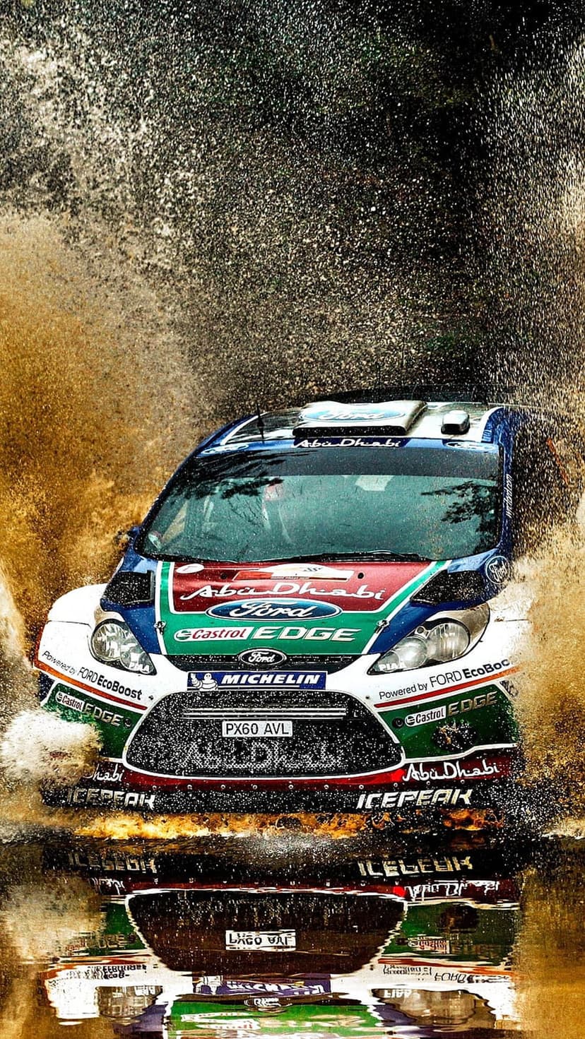Ford rally car kicking up a massive spray of muddy water while racing on a dirt track with sponsor logos visible.