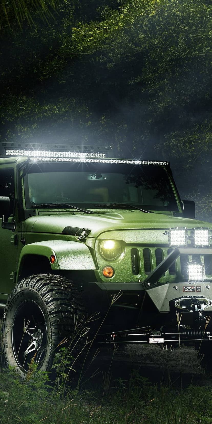 Green Jeep Wrangler with glowing LED light bars parked in a misty forest at night with large off-road tires.