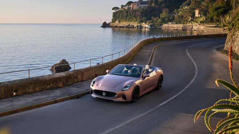 Maserati GranCabrio convertible cruises along a winding coastal highway next to bright blue ocean water under clear sky.