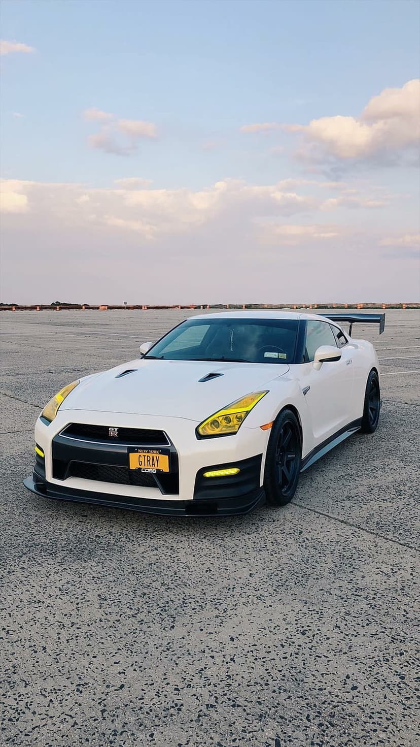 Nissan GT-R in white with yellow headlights and GTRAY license plate on asphalt under a cloudy blue sky.