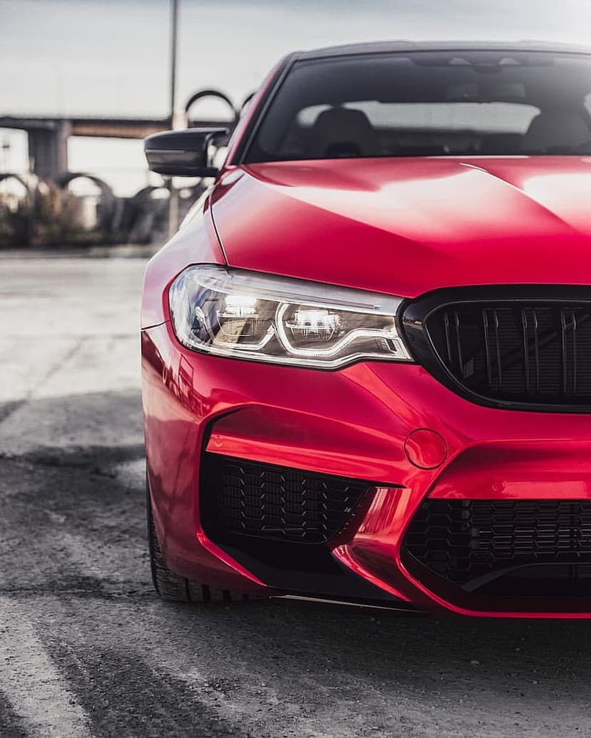 Red BMW M5 sedan front view with glowing LED headlights and a black kidney grille against an urban backdrop.