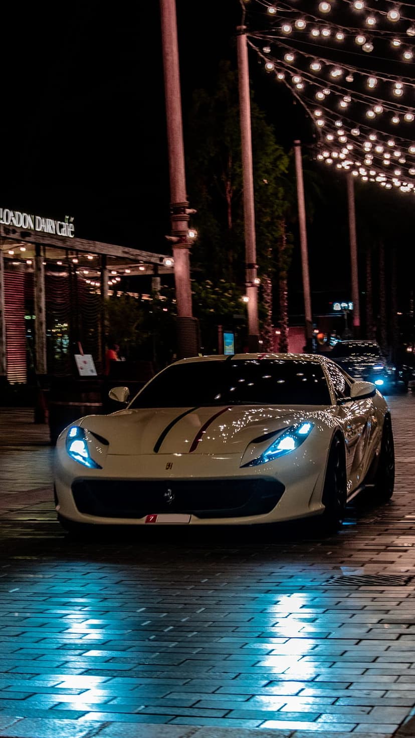 White Ferrari supercar with blue headlights and red stripes parked on a wet brick city street under night lights.