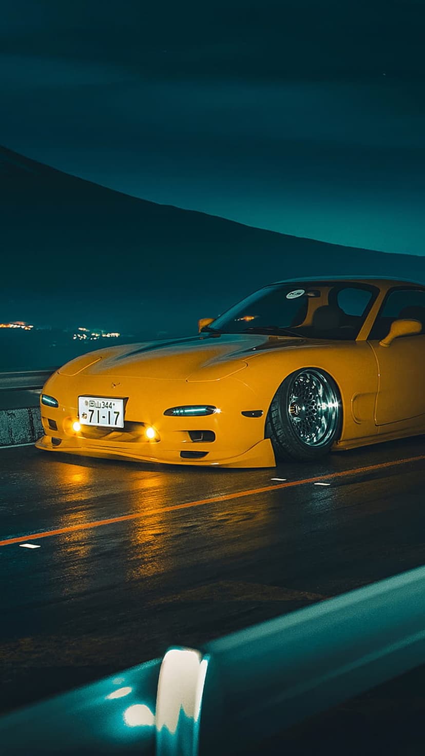 Yellow Mazda RX-7 with glowing headlights on a wet road at night under a dark teal sky and mountain backdrop.