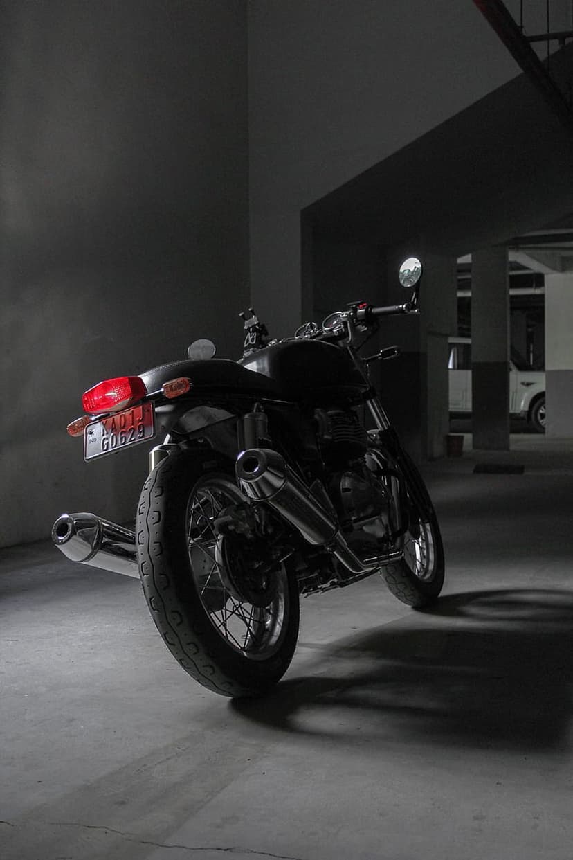 Royal Enfield motorcycle with twin exhaust pipes and a red taillight parked in a dark, shadowy indoor garage.