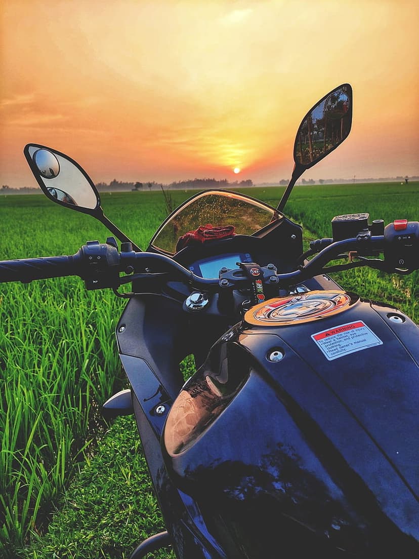Motorcycle parked in a green field reflecting orange sunset light on its windshield and metallic fuel tank.