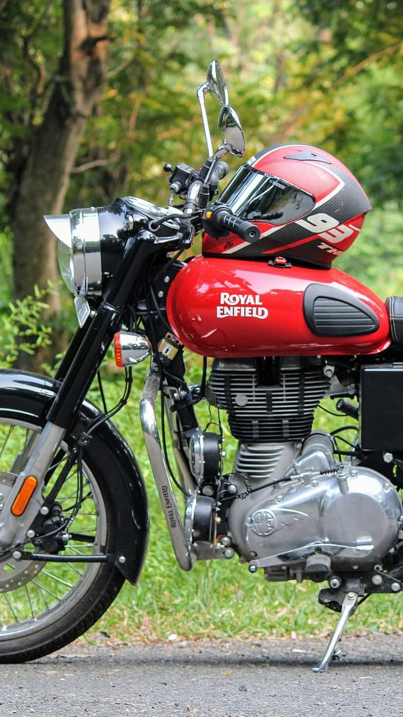 Red Royal Enfield motorcycle with a helmet on the tank parked in a green forest with visible chrome engine parts.