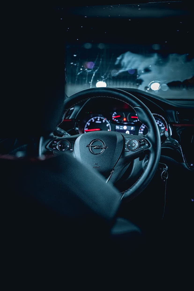 Steering wheel and glowing dashboard dials overlook blurred city lights through a car windshield at night.