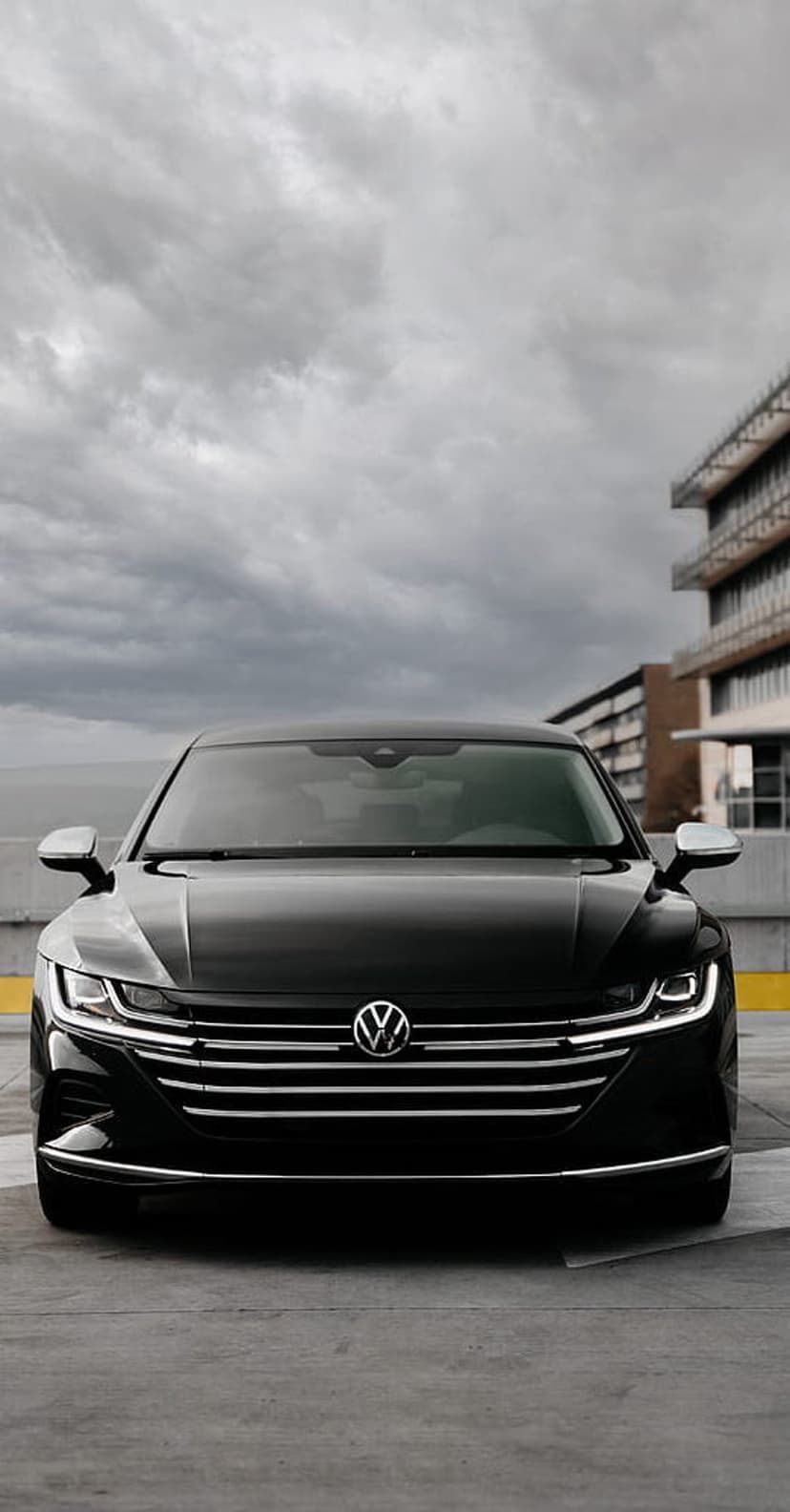 Black Volkswagen Arteon front view with LED headlights and chrome grille against a cloudy urban background.