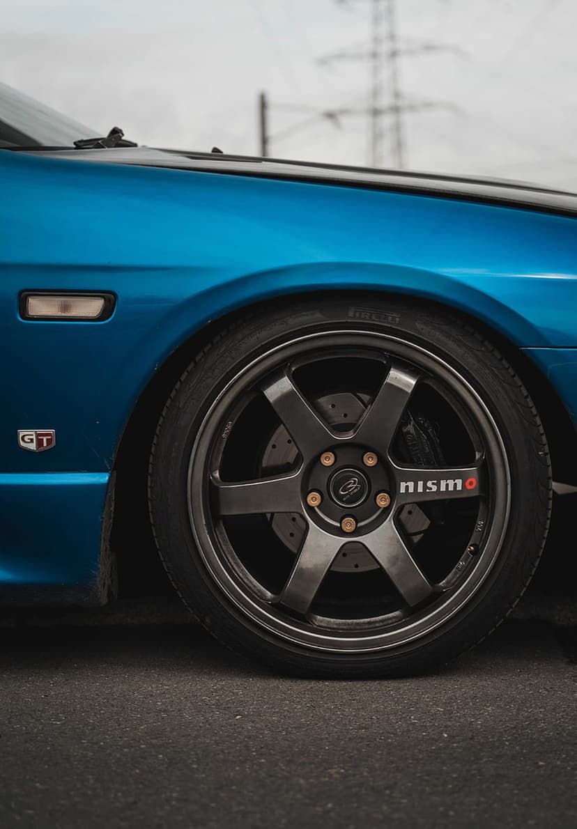 Nissan GT-R R33 blue fender with a dark grey Nismo wheel, gold lug nuts, and a silver GT badge in a close-up shot.