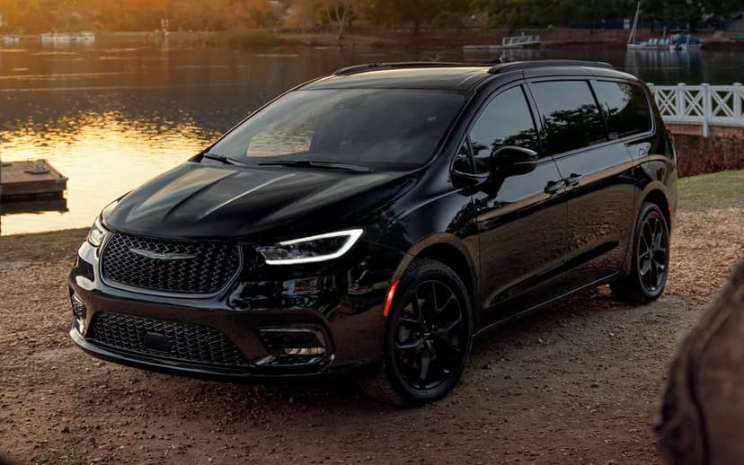 Black Chrysler Pacifica minivan parked on a dirt path beside a lake at sunset with golden reflections.