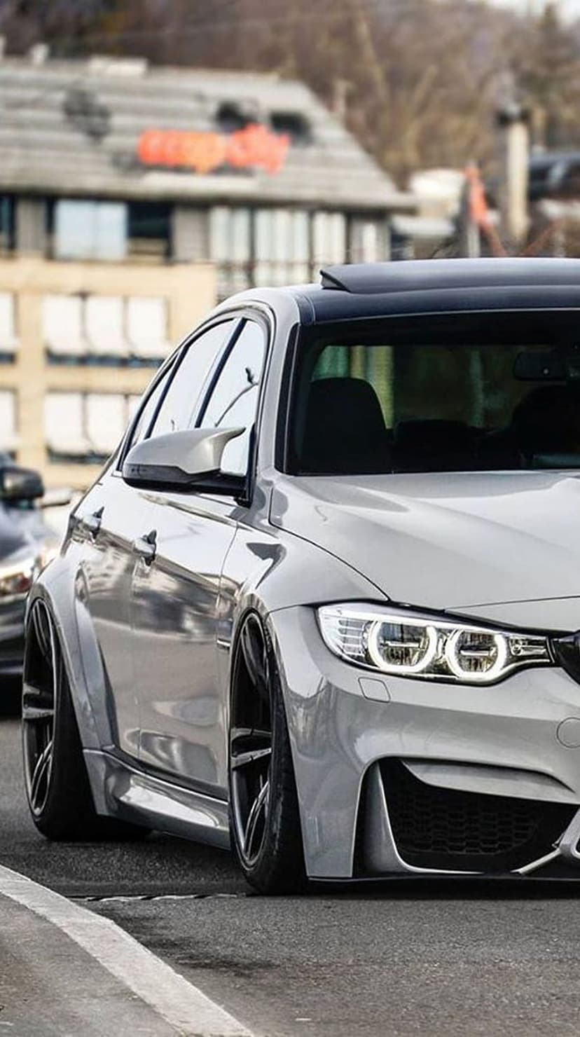 BMW M4 in slate grey features black multi-spoke rims, a widebody kit, and a slammed stance on a city street.