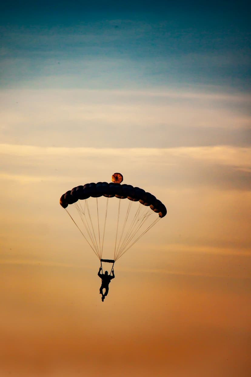 Skydiver silhouetted against a bright orange and yellow sunset sky while descending under an open parachute.