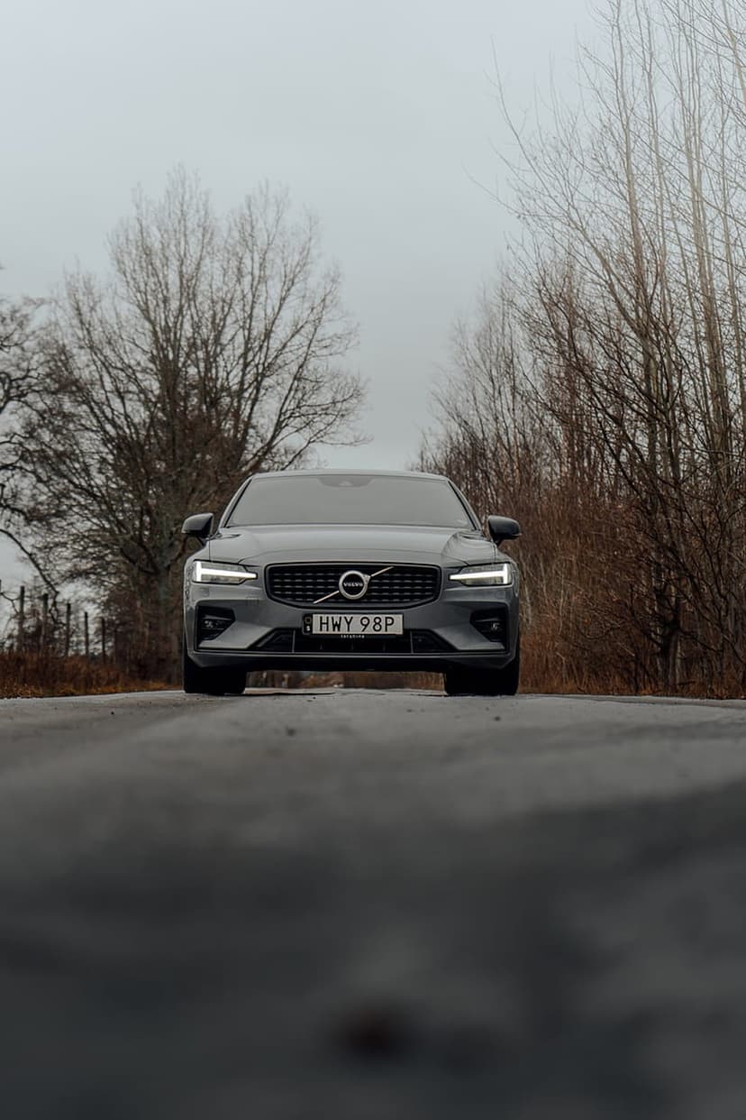Volvo S60 sedan with glowing headlights parked on a wet, foggy road surrounded by bare winter trees in the mist.