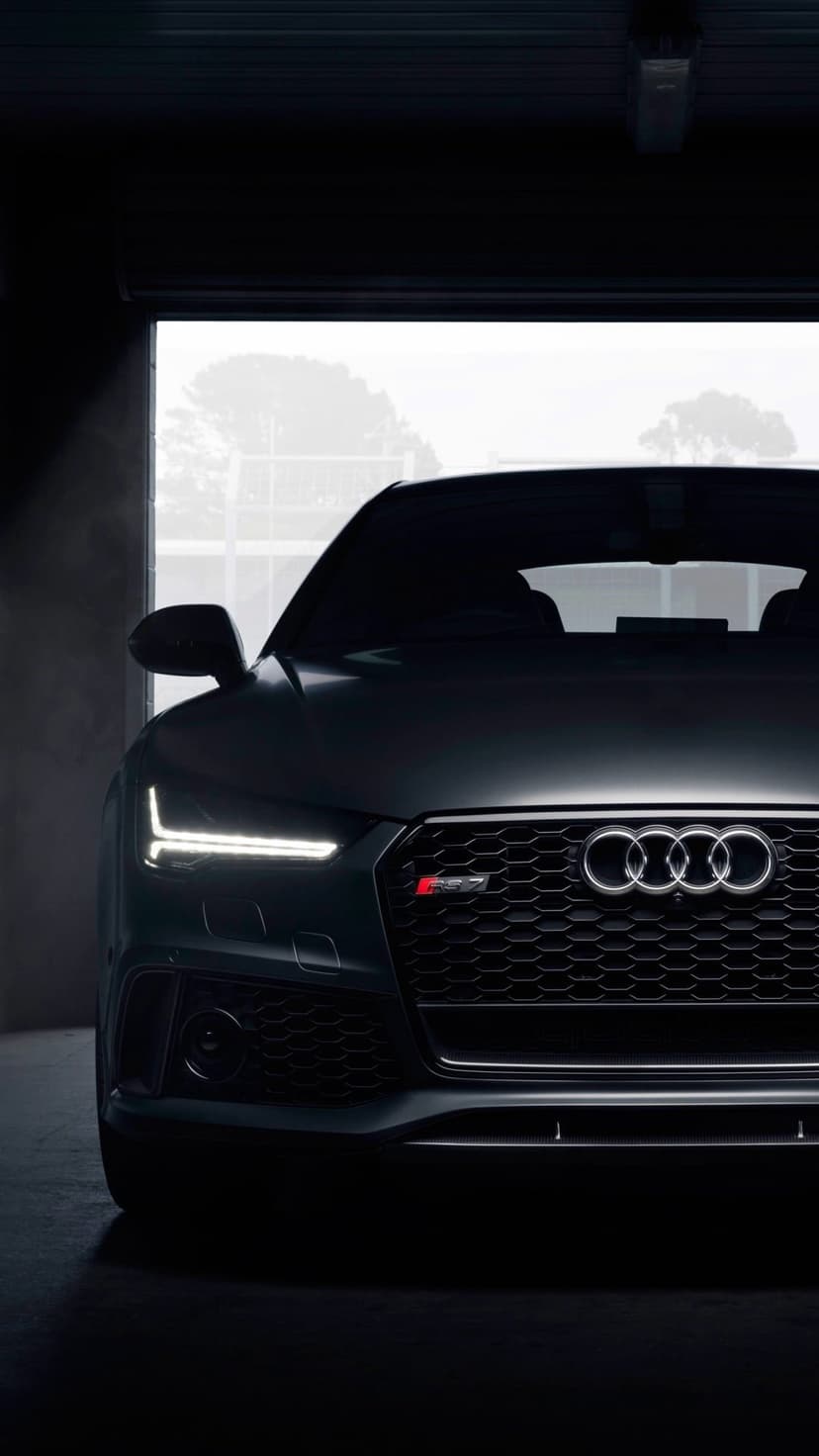 Audi RS7 sports car with glowing headlights parked in a dark garage revealing a glimpse of trees behind.