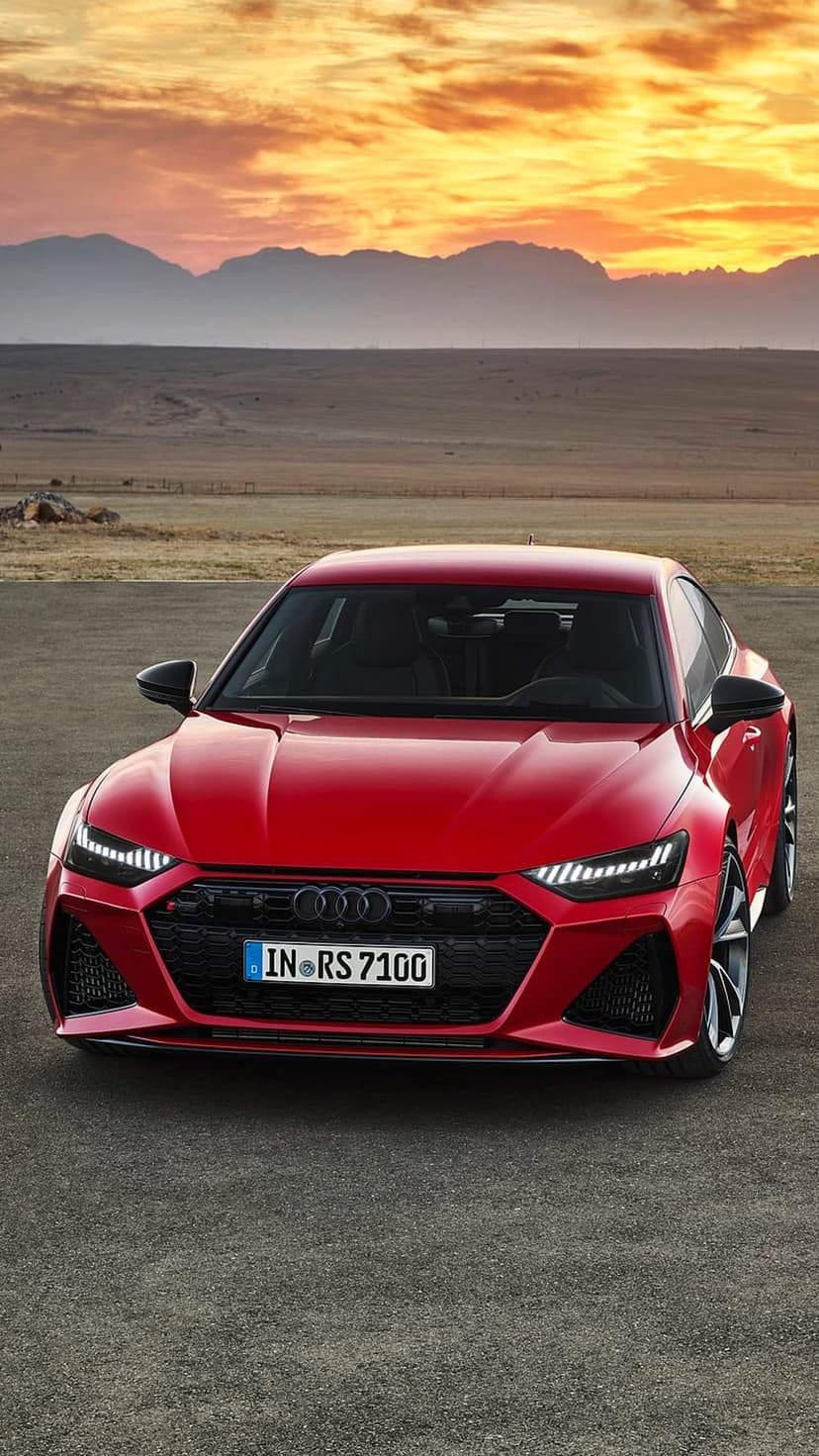 Red Audi RS7 Sportback parked on a desert road under a glowing orange sunset sky with mountain silhouettes.
