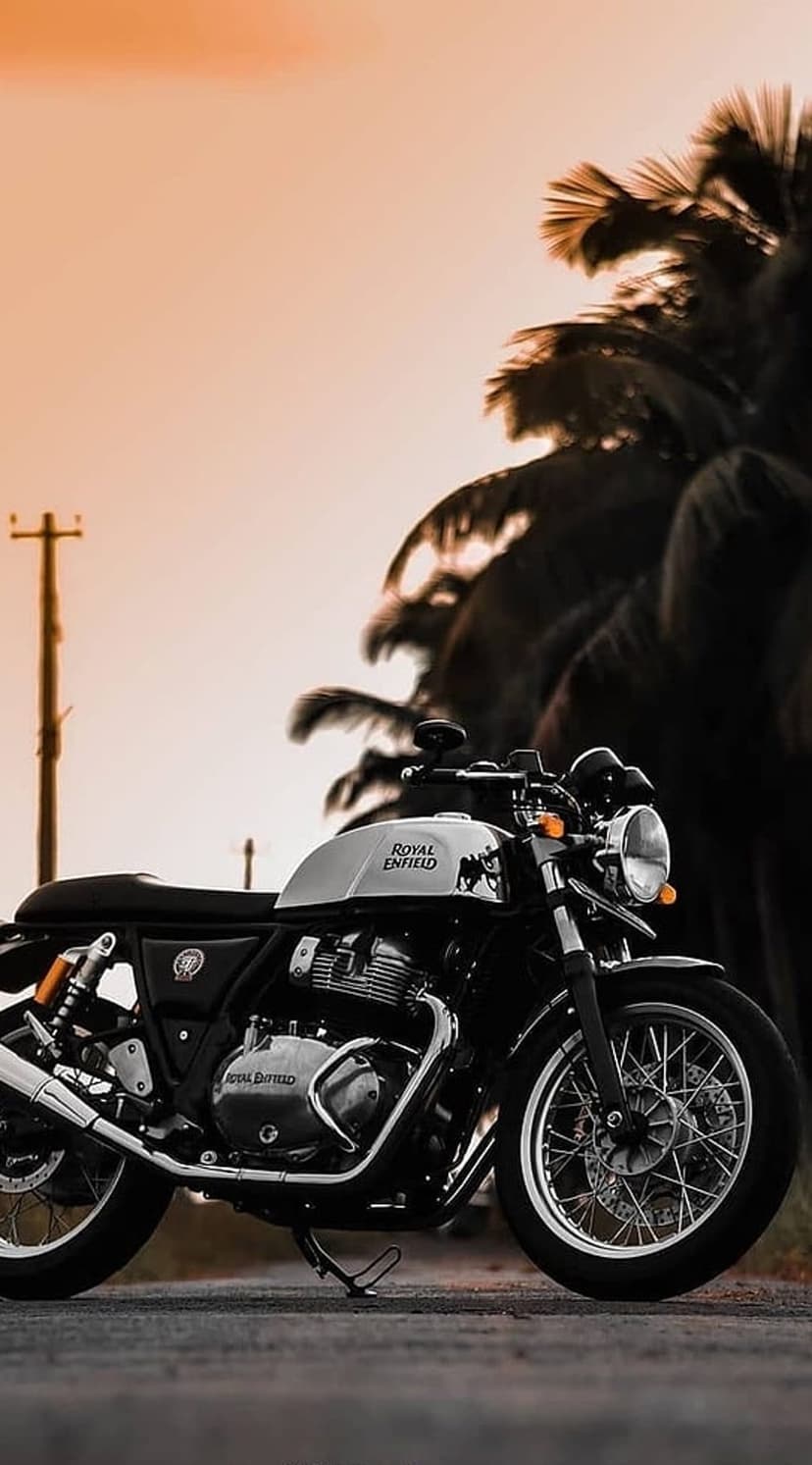 Royal Enfield motorcycle parked near silhouetted palm trees under a glowing orange and yellow sunset sky.