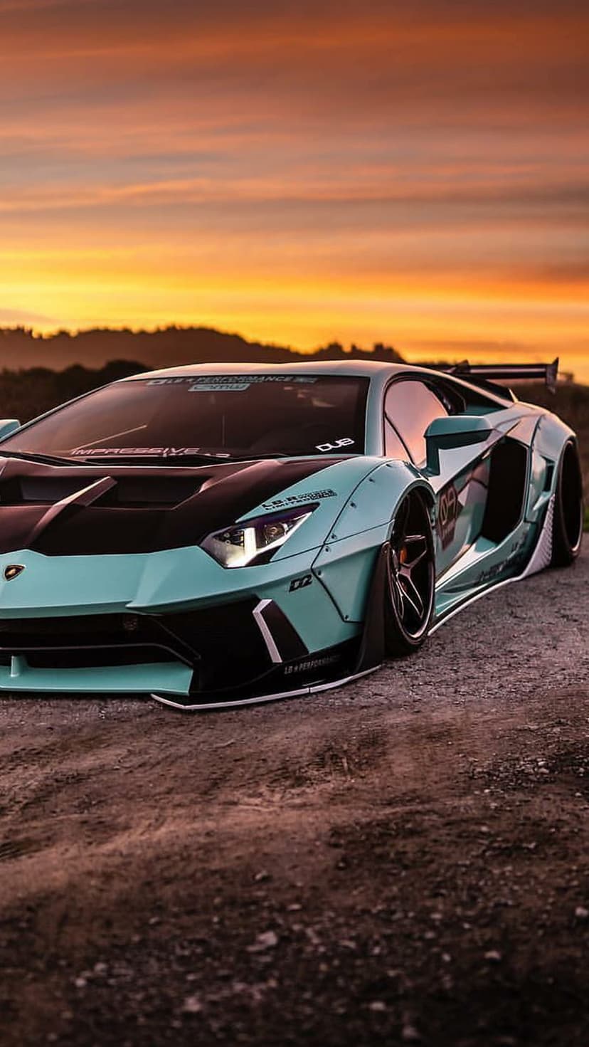 Cyan Lamborghini Aventador with widebody kit parked on a dirt road during a warm orange and purple sunset sky.
