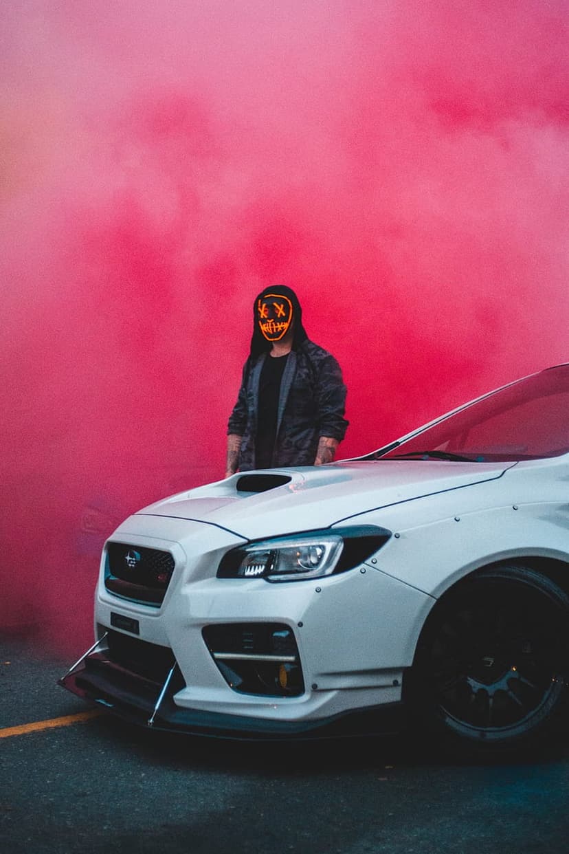 Person in a glowing orange mask stands by a white wide-body Subaru surrounded by thick vibrant pink smoke.