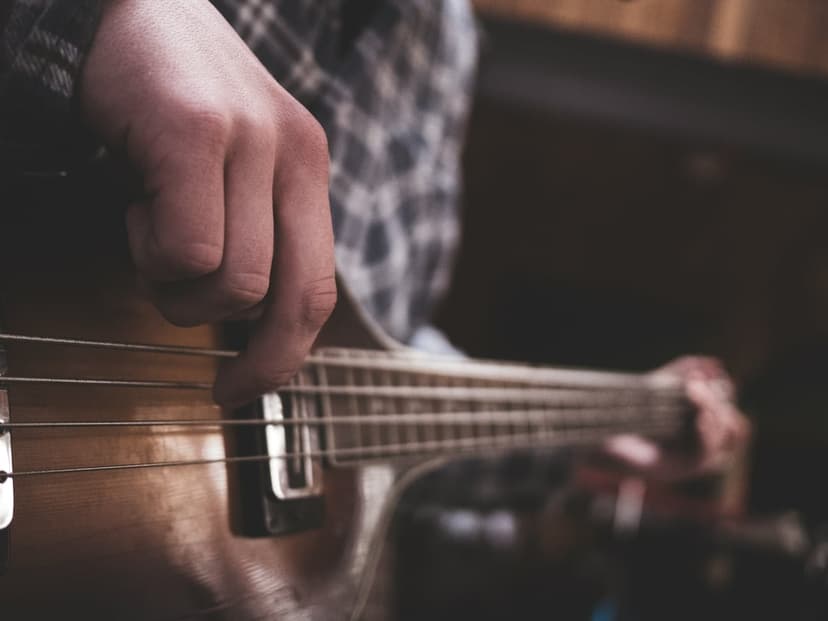 Musician's hand strums thick metal strings on a polished wood bass guitar with a blurred plaid shirt sleeve.
