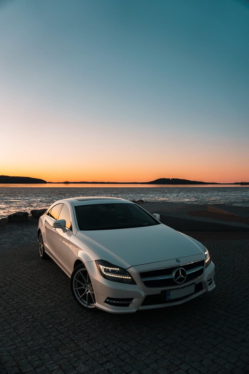 Mercedes CLS sedan with glowing headlights on a rocky cobblestone beach against a vibrant orange sunset.