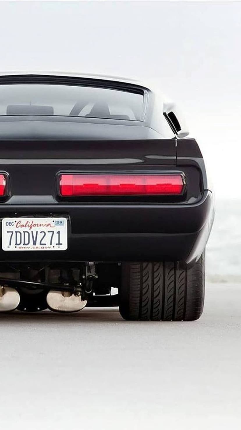 Black Dodge Challenger rear with red taillights and California plate on a beach with a blurred ocean backdrop.