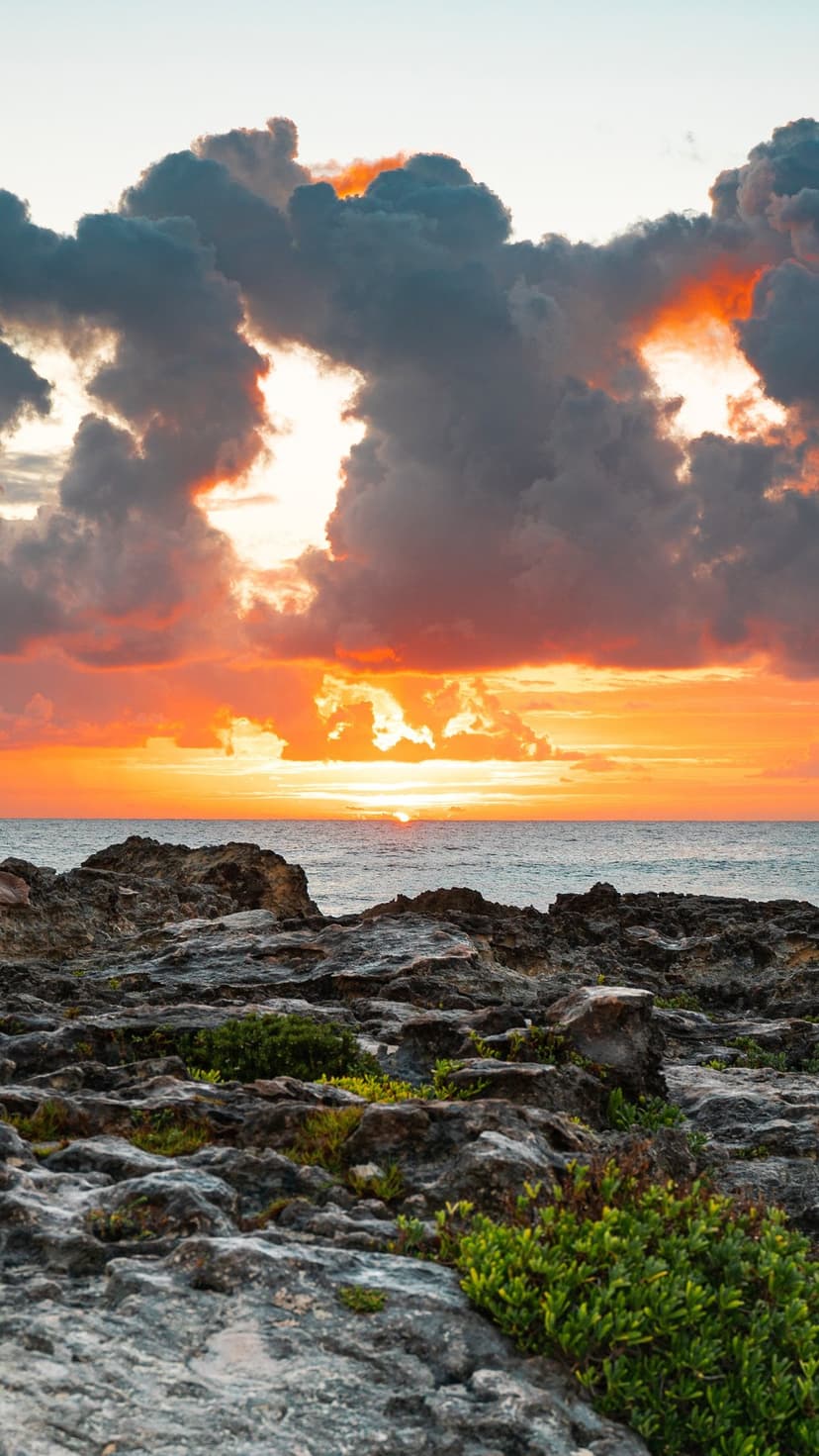Vivid orange sunset sky with dark clouds overlooking a jagged rocky shoreline and calm ocean waters at dusk.