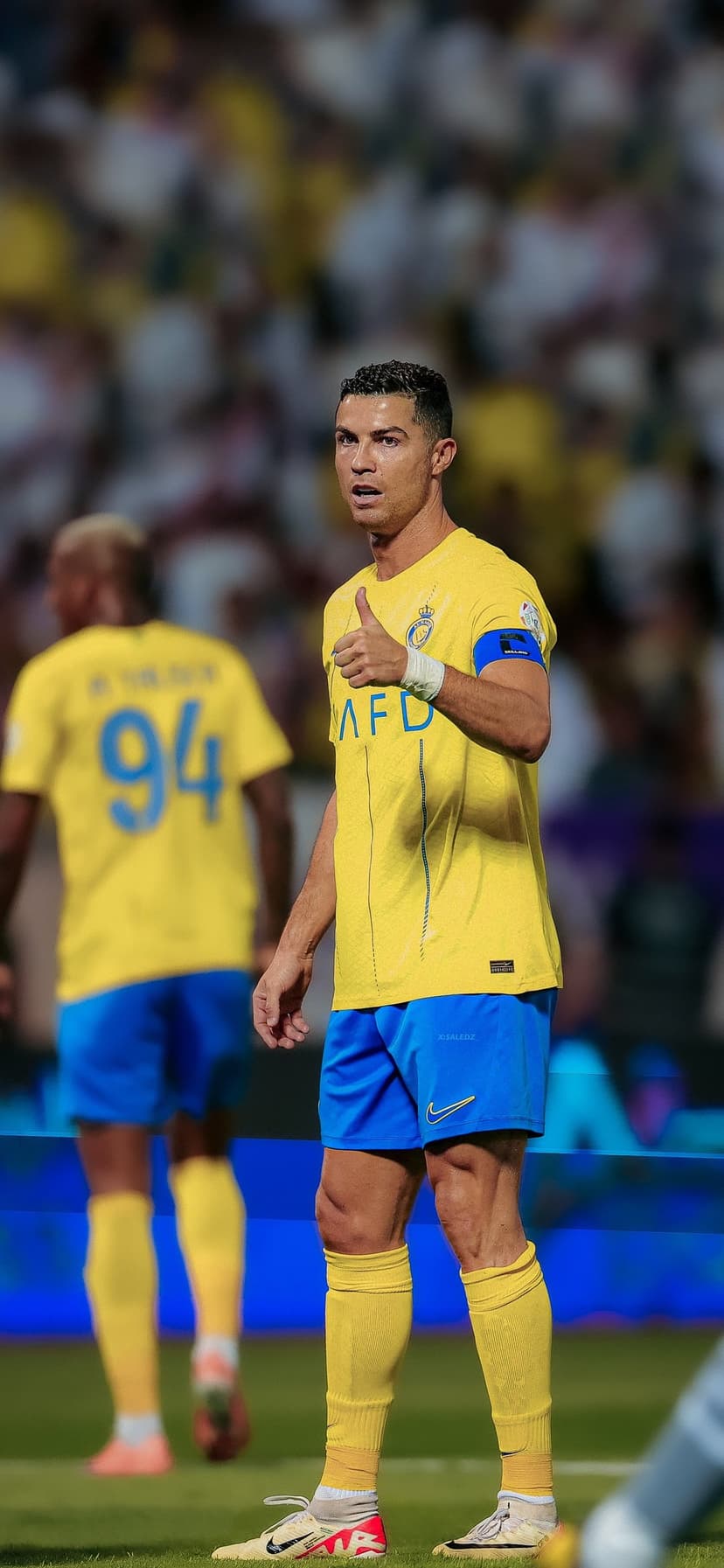 Cristiano Ronaldo gives a thumbs up while wearing a yellow and blue Al Nassr jersey on a green soccer field.