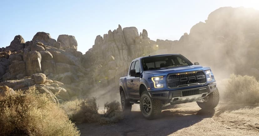 Blue Ford F-150 Raptor truck driving through a dusty desert landscape with large rock formations and sun flare.
