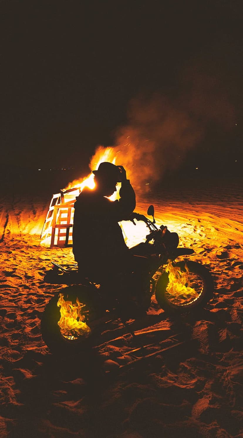Motorcyclist in a hat sits on a bike before a massive beach bonfire with soaring orange flames and thick smoke.