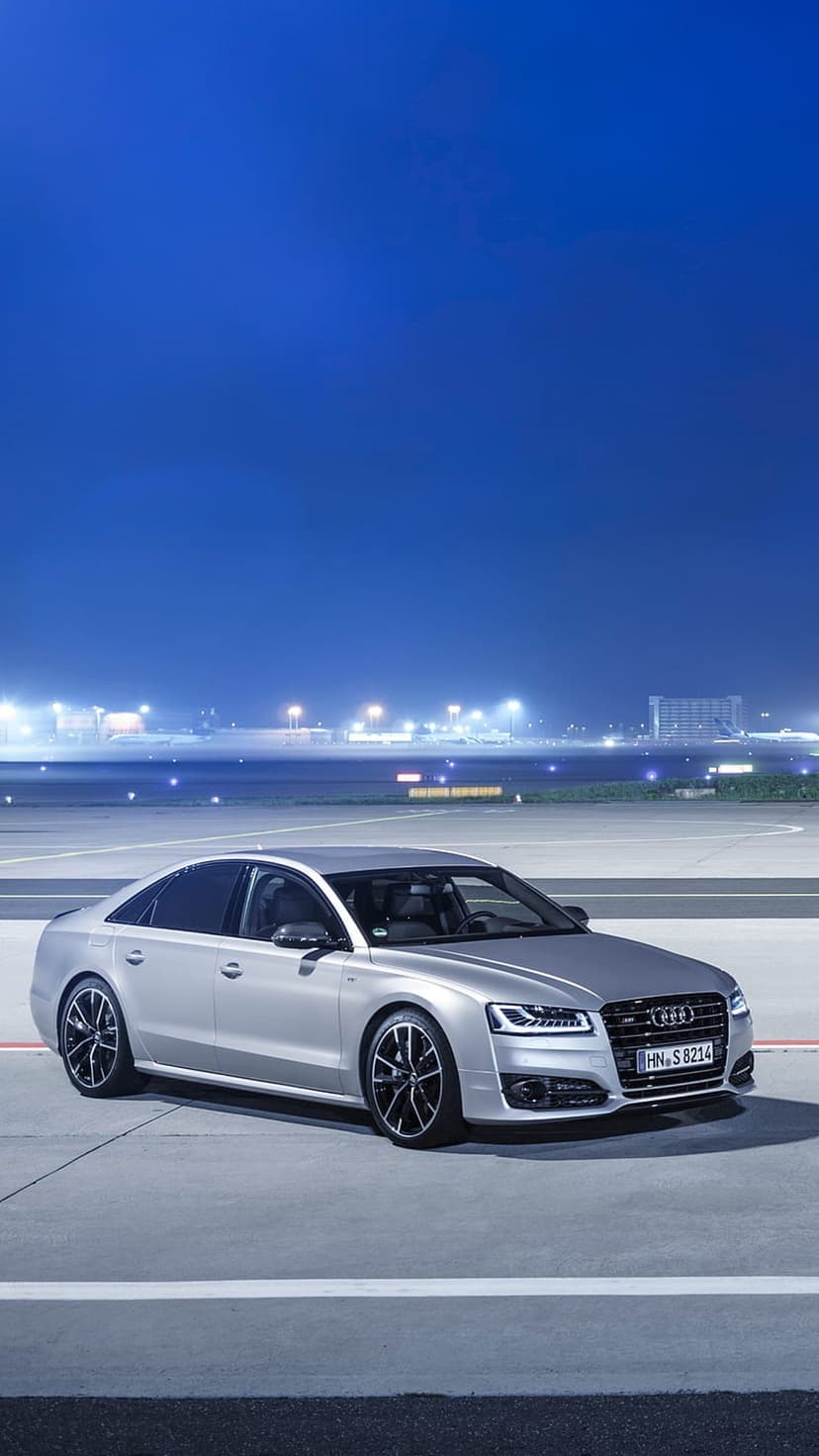 Silver Audi S8 luxury sedan with glowing headlights parked on a dark airport tarmac under a blue night sky.