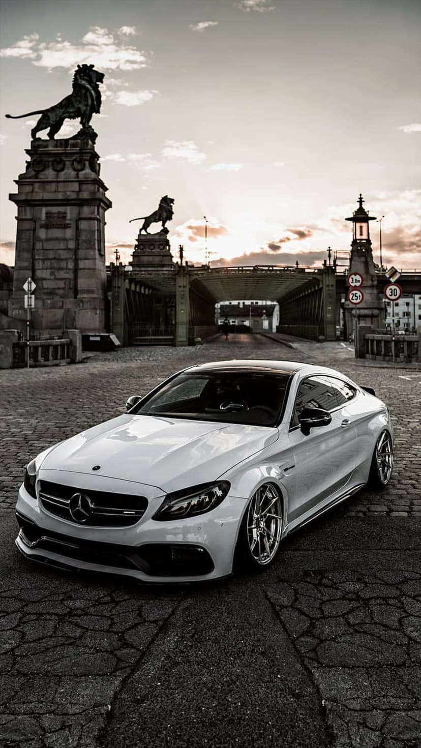 White Mercedes C63s Coupe parked on cobblestones by an arched bridge with lion statues under a sunset sky.