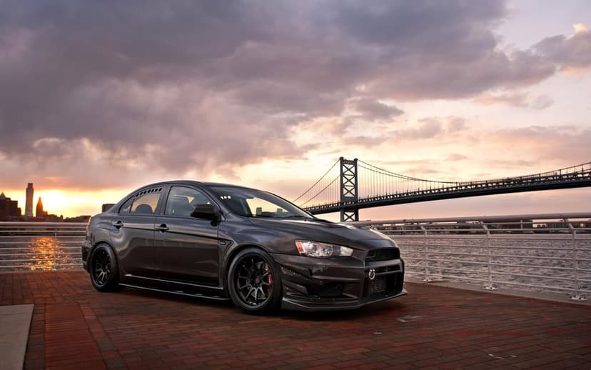 Dark grey Mitsubishi Lancer Evo X parked on a brick pier with a suspension bridge under a sunset sky.