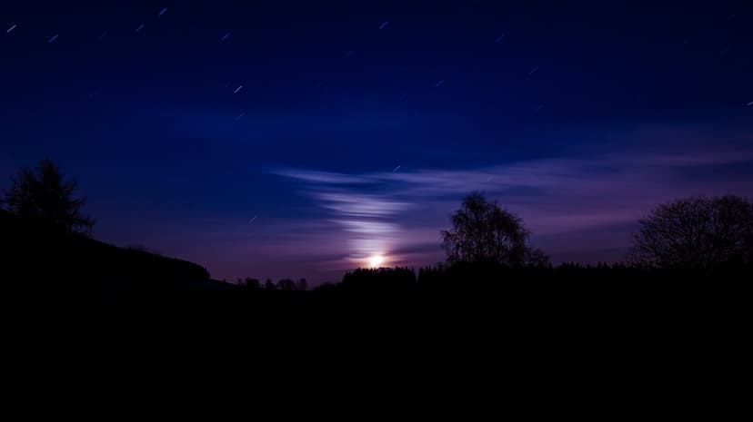 Luminous moon peeking through thin clouds above dark silhouettes of forest trees and rolling hills at night.