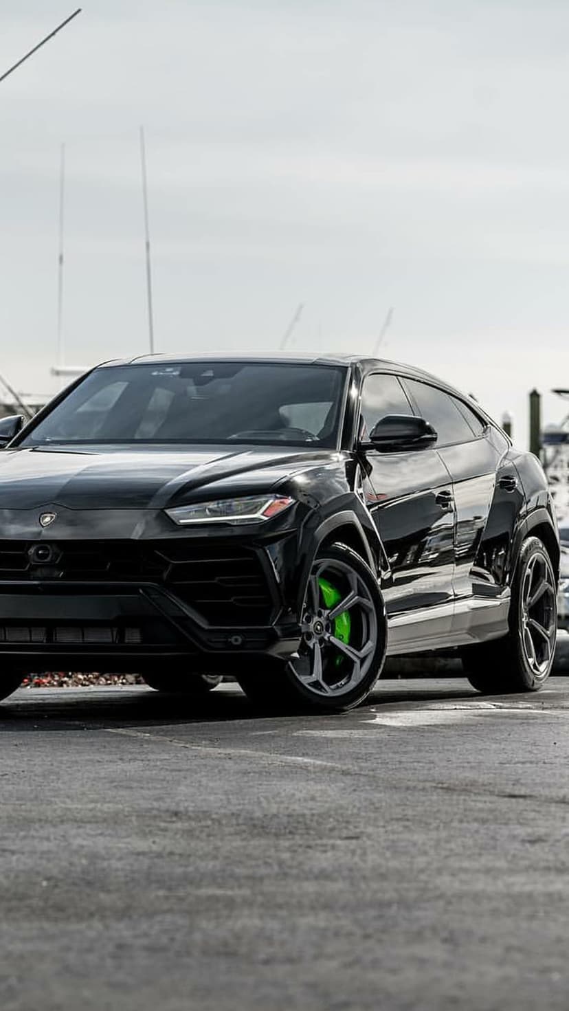 Black Lamborghini Urus SUV with neon green brake calipers parked on pavement near water in a vertical portrait.