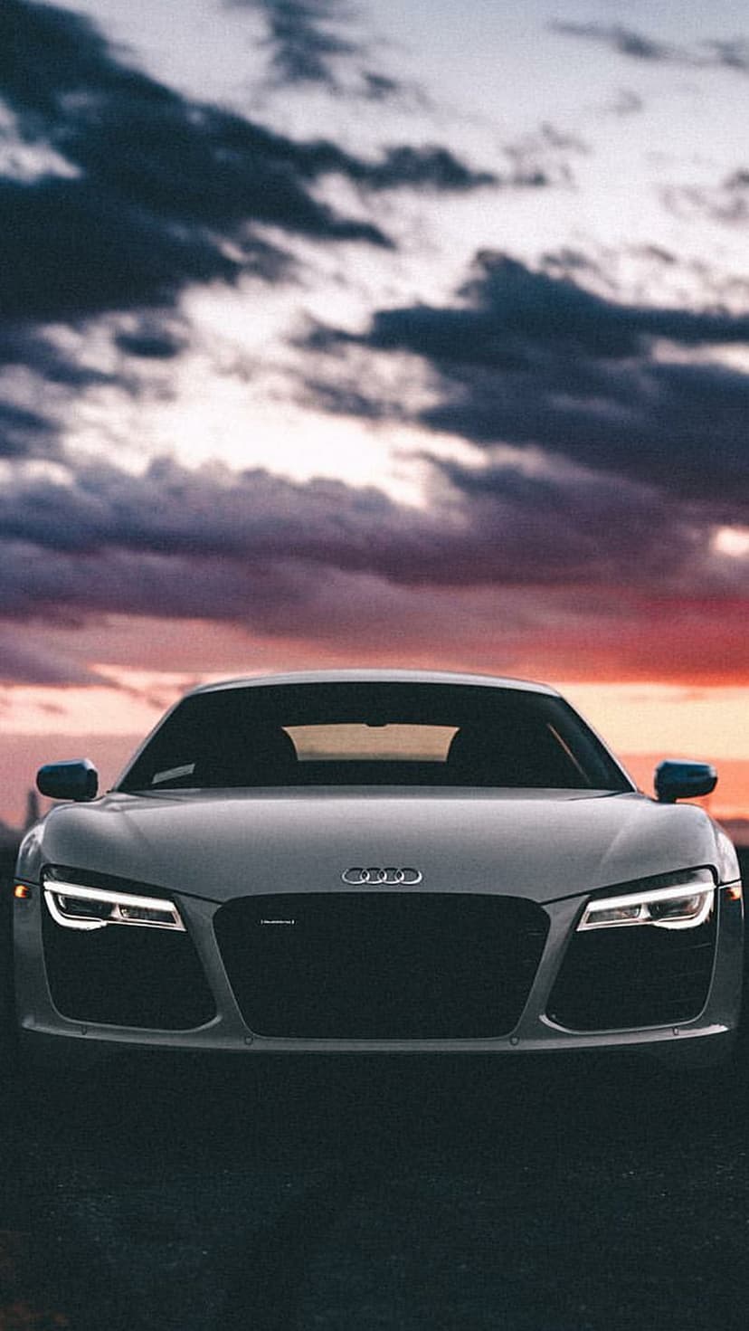 Audi R8 sports car with illuminated headlights parked under a dramatic orange and dark grey sunset sky.