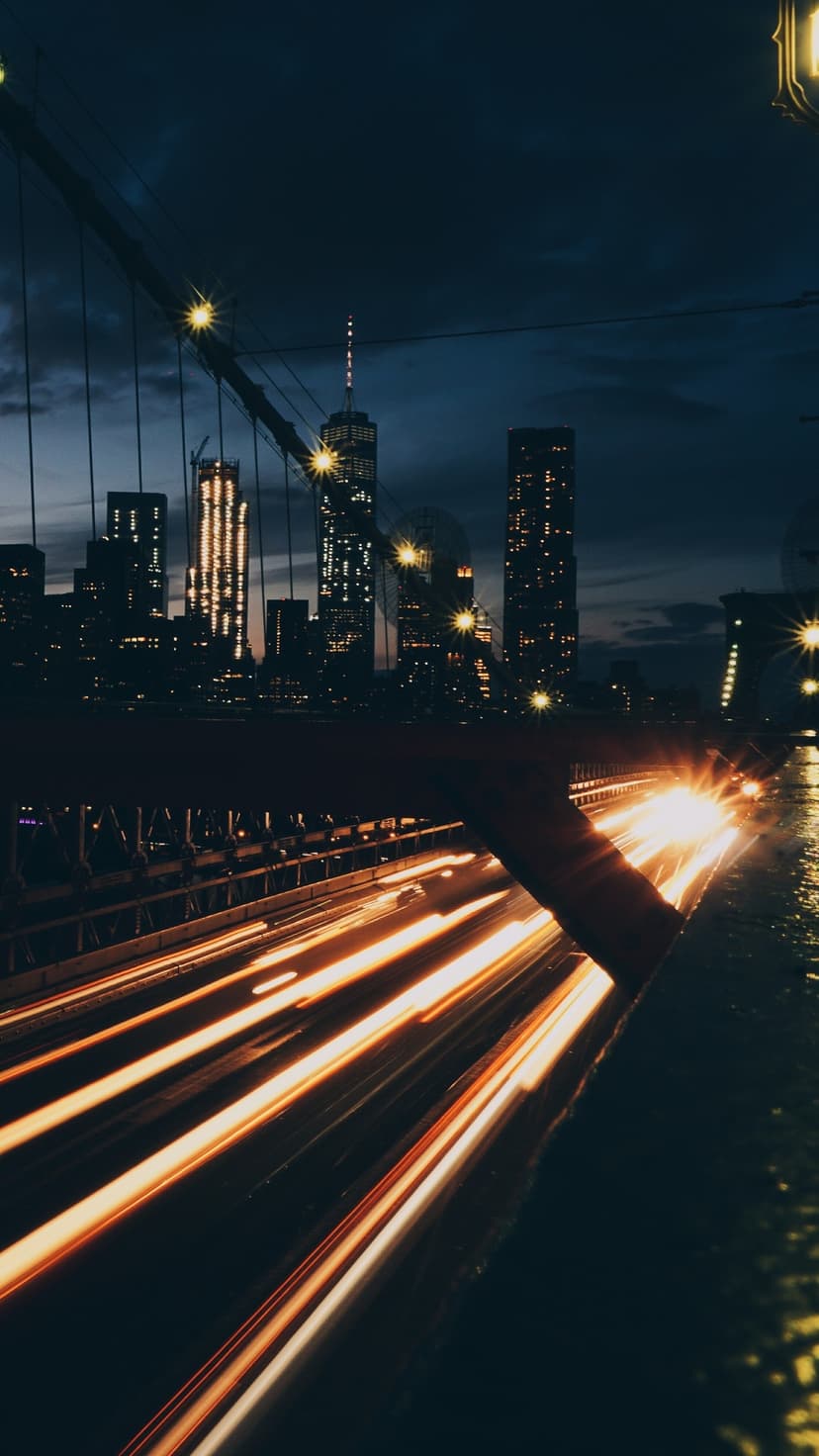 Golden traffic light trails on the Brooklyn Bridge with the glowing Manhattan skyline under a dark cloudy sky.
