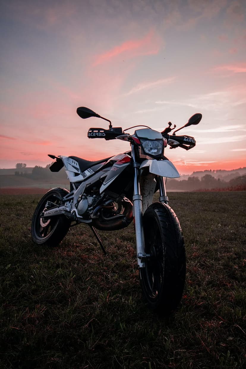 Supermoto motorcycle parked in tall grass under a vibrant orange and pink sunset sky with soft lighting.