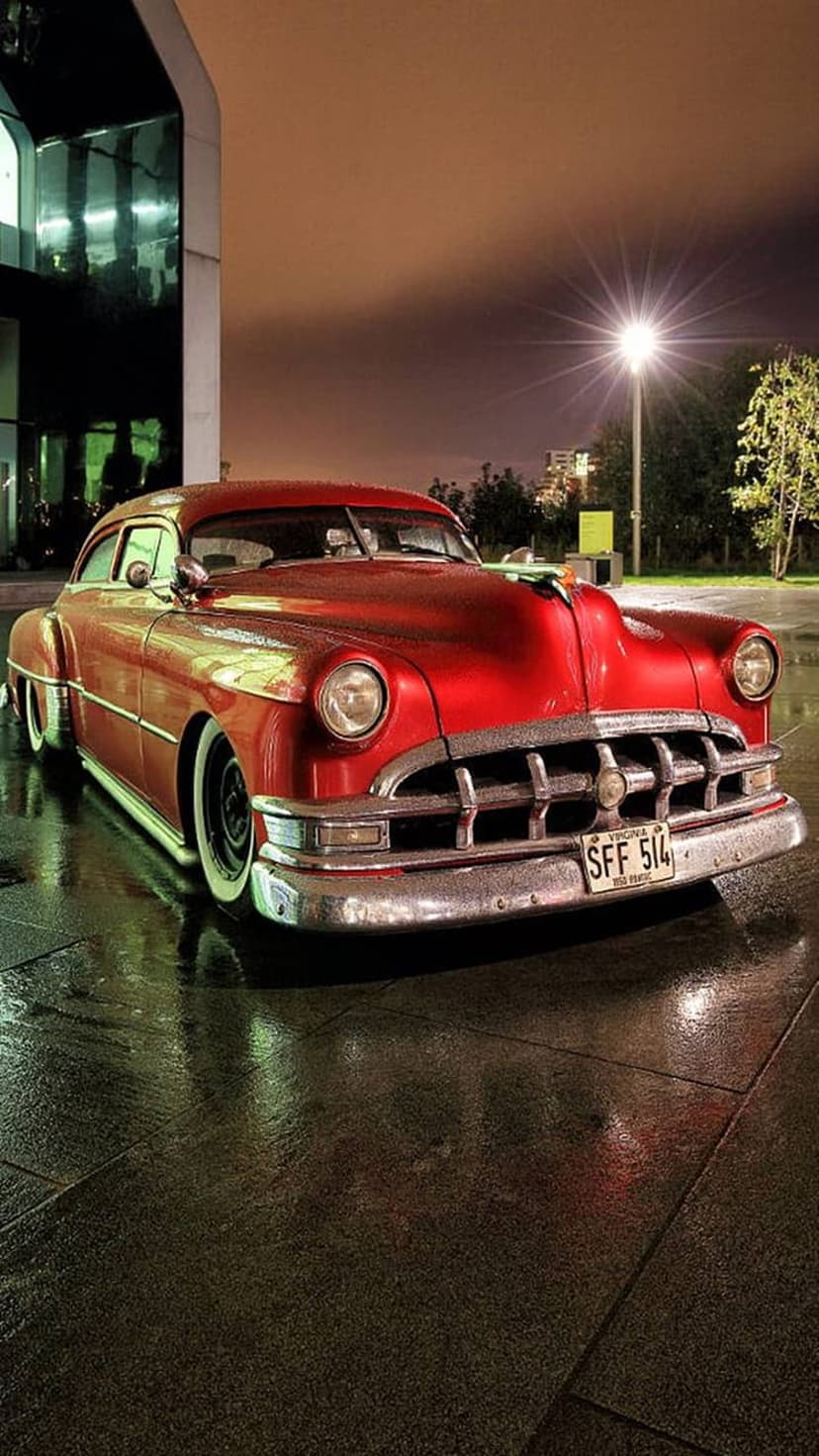 Crimson vintage car with chrome details rests on wet, reflective pavement under a night streetlamp's glow.