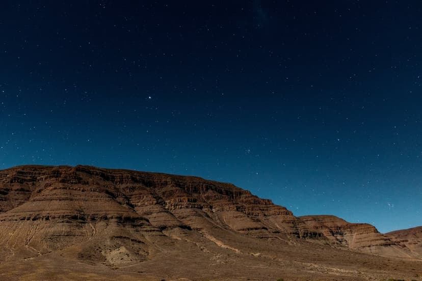 Stratified desert mountains rise toward a clear midnight sky illuminated by countless twinkling stars and nebulae.