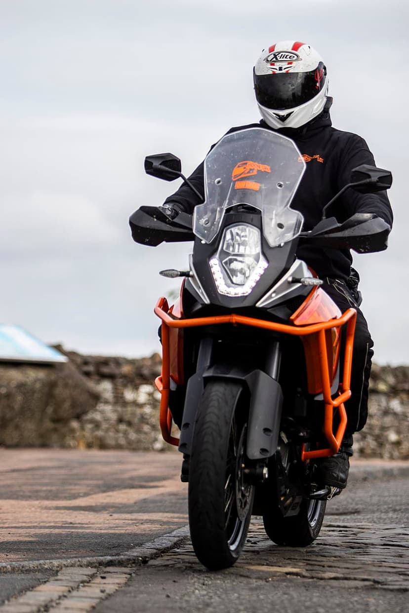 Motorcyclist in a red and white helmet rides a black and orange adventure bike past a grey stone wall.