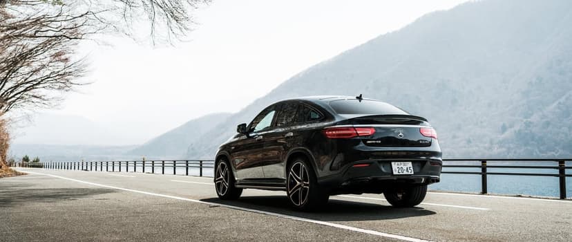 Rear view of a black Mercedes-AMG GLE 43 Coupe on a mountain road beside a lake with hazy peaks in the background.