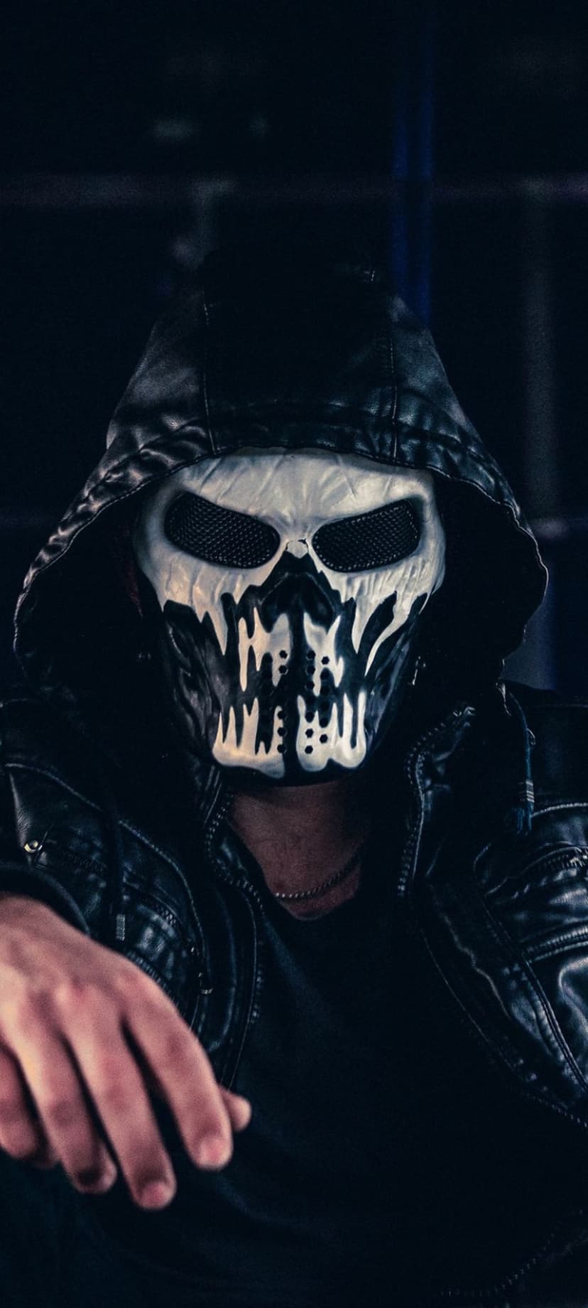 Person in a white skull mask and black leather hooded jacket stares forward against a dark, moody background.